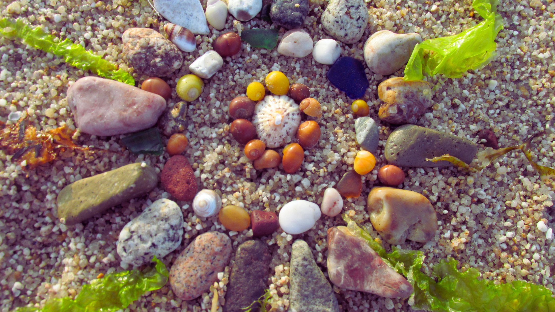 Mandala de coquillages, galets et verre de mer disposés en cercles sur le sable, ambiance dorée